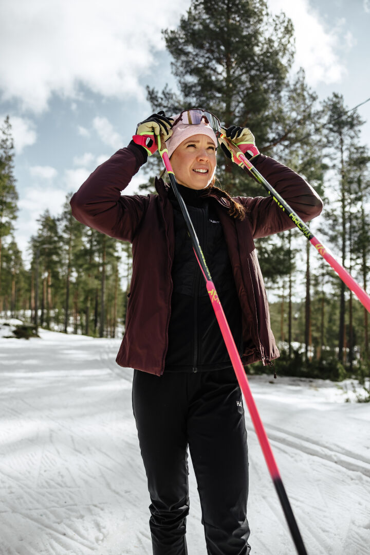 A woman in a Halti Haave Primaloft Insulation Jacket smiles, holding ski poles on a snowy forest trail.