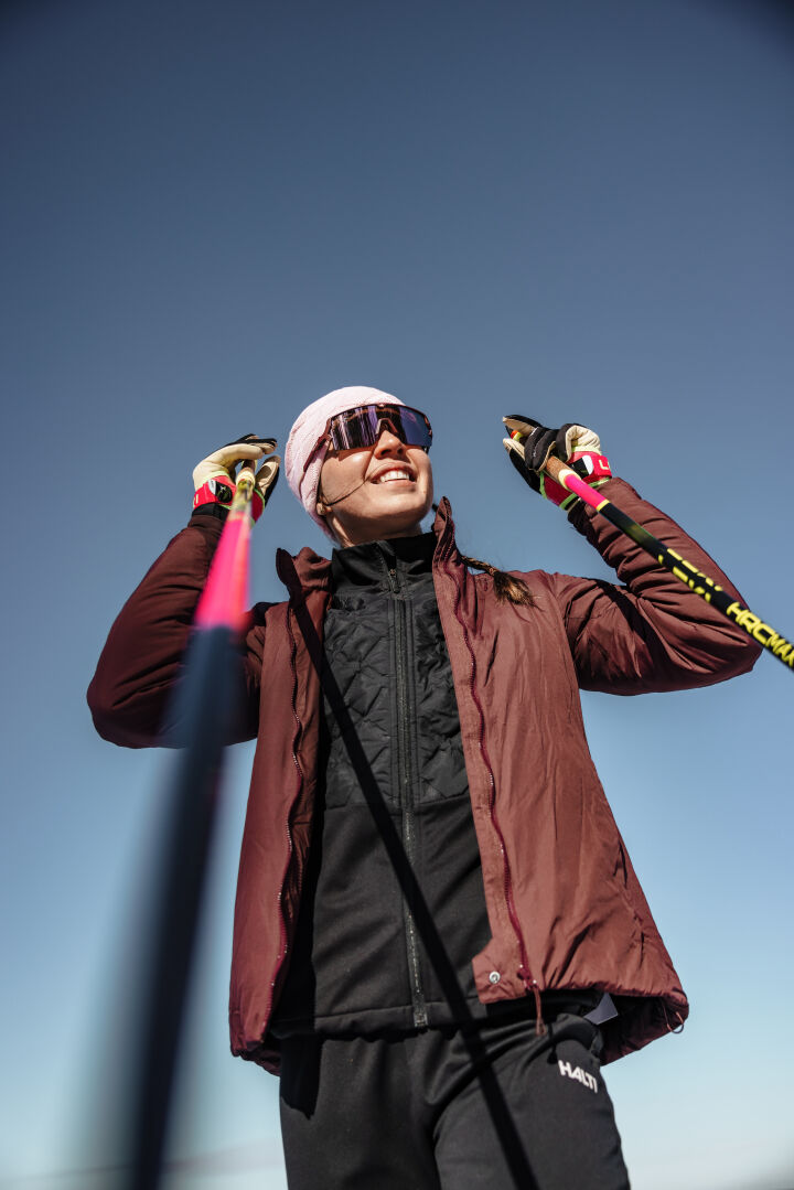 A woman in a Halti Haave PrimaLoft Insulation Jacket and sunglasses smiles, holding ski poles under a clear blue sky.