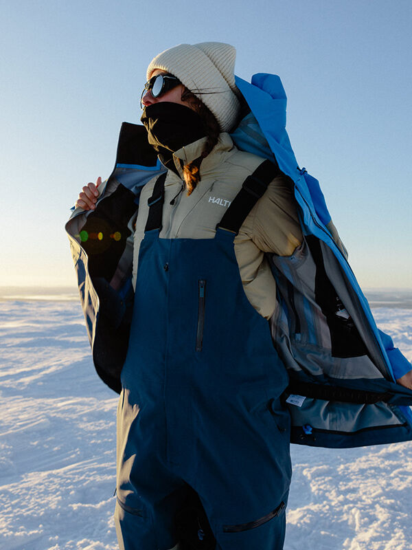 A person in winter gear stands on snow, wearing sunglasses, a beanie, and the Halti Haave Primaloft Insulation Jacket Women's.