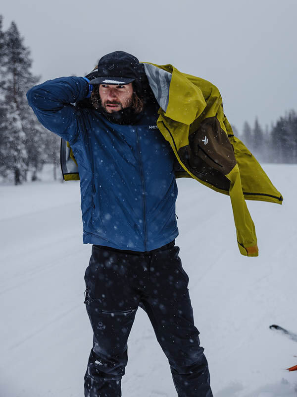Wearing the Halti Rizle 3L DX Ski Jacket Unisex, a person stands outdoors in snow with trees behind.