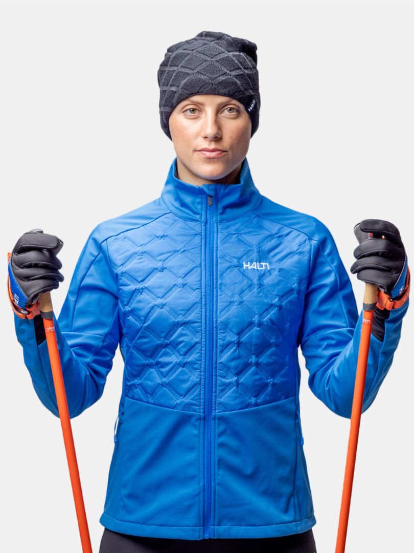 Wearing a Halti Crostie Merino Beanie, a person in blue with orange ski poles faces forward on a white background.