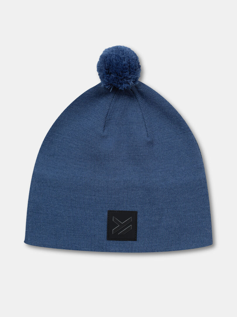 The Halti Pilke Beanie is a blue merino knit with pom-pom and black patch—ideal for winter sports or skiing.