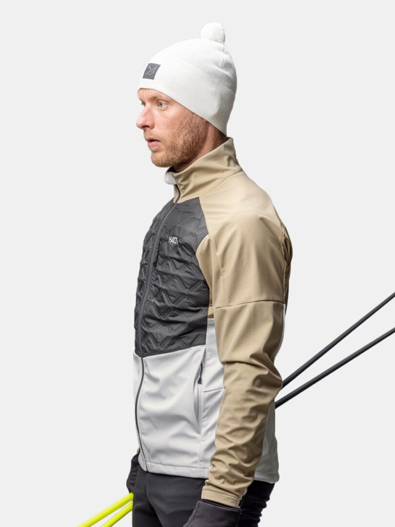 A man in a beige, gray, and black jacket wears the Halti Pilke Beanie, holding ski poles against a plain background.