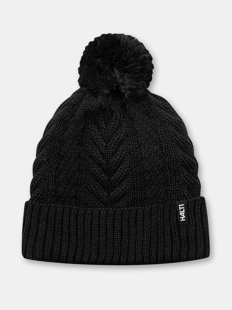 Halti Hile Beanie: black cable knit, fleece-lined, pom-pom top, and branded tag on the folded brim.