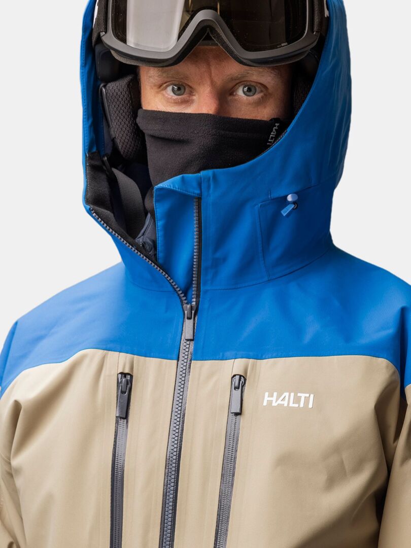Person in a Halti Trone Tube Scarf Fleece, blue and beige Halti jacket, and ski goggles—styled for cold weather.