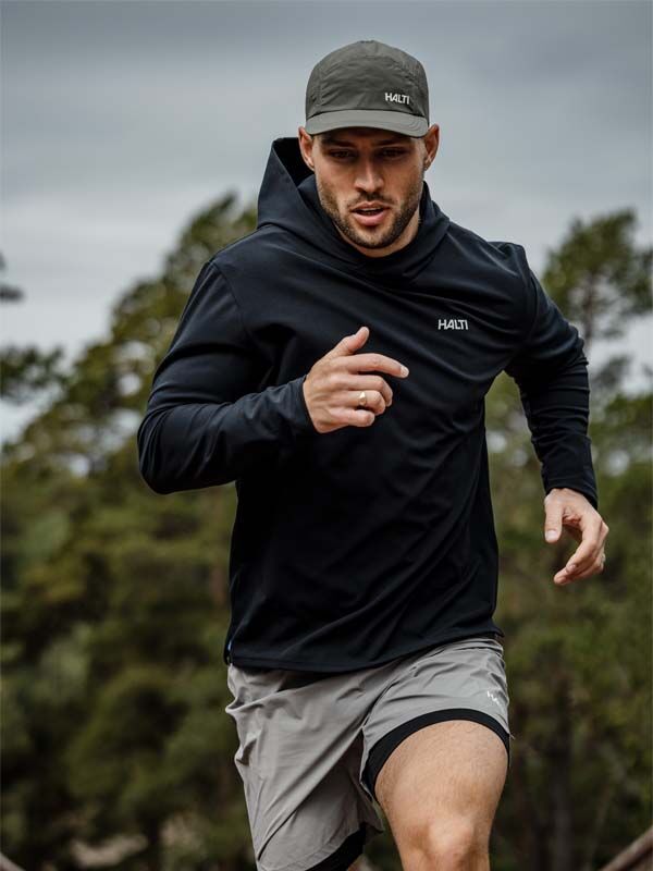 A man in Halti Uurto Shorts Men's and a black hoodie runs outdoors with trees and a cloudy sky in the background.