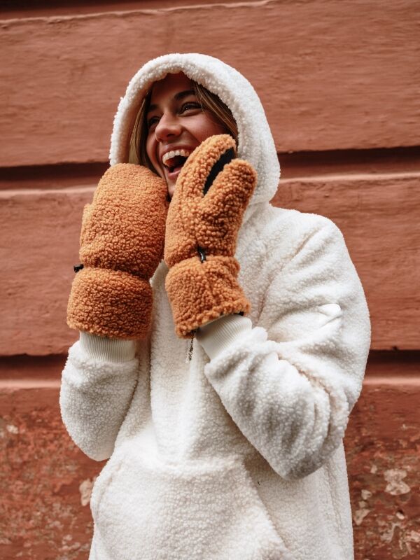 Someone wearing Halti’s Lykky Fleece Mittens and a white hooded jacket smiles in front of a textured red wall.
