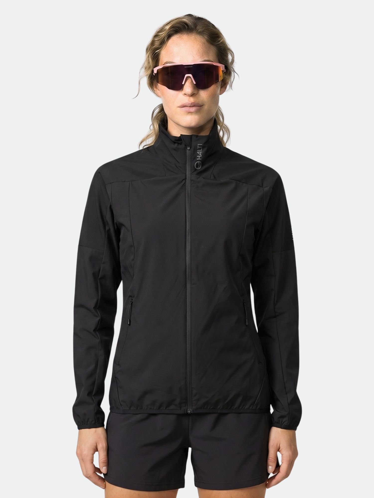 A person wears the Halti Urbanite Women's Training Jacket in black with matching shorts and sunglasses against a light background.
