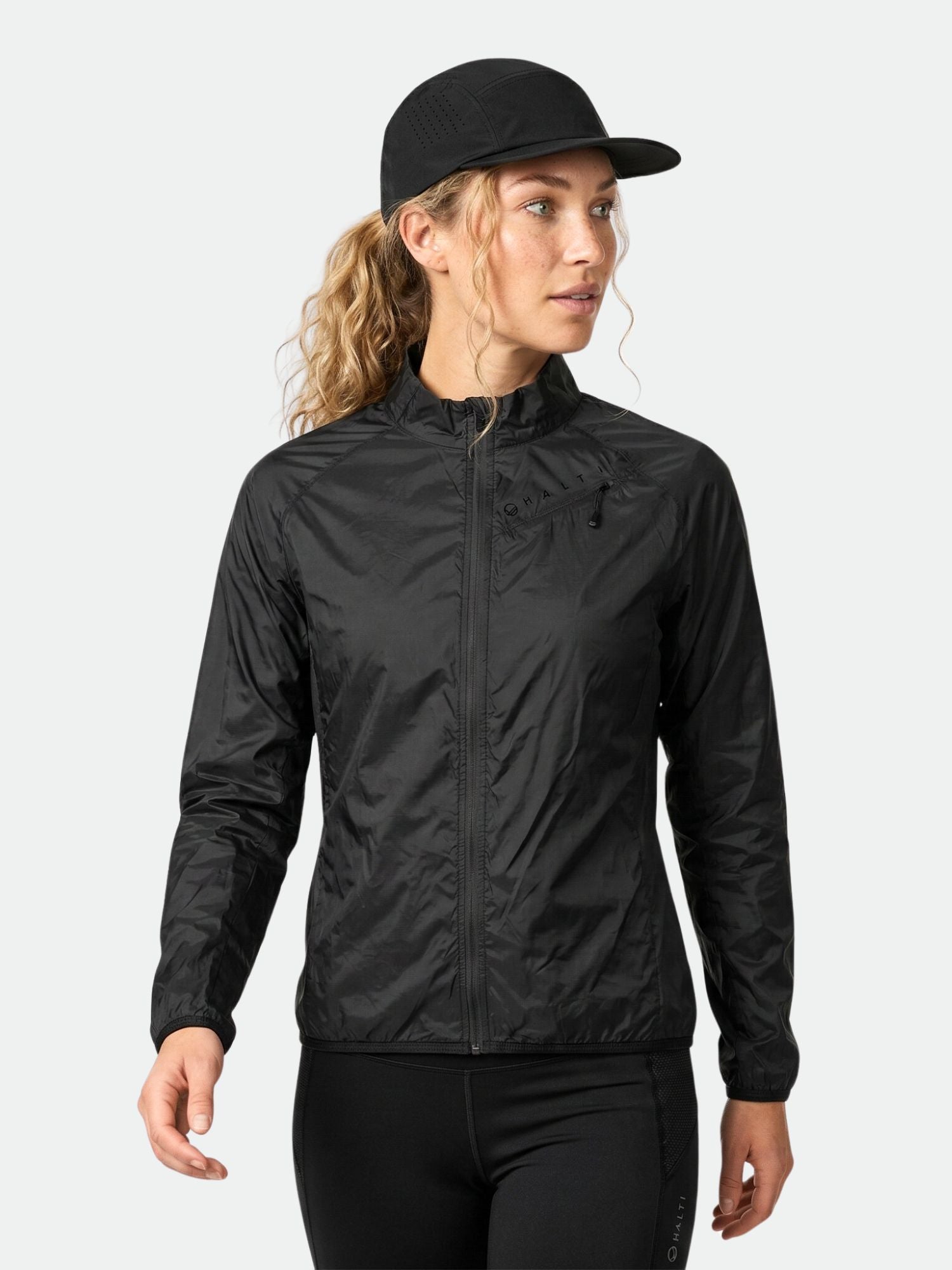 A woman wears the Halti Control Women's Windbreaker Jacket, black pants, and a black cap against a white background.