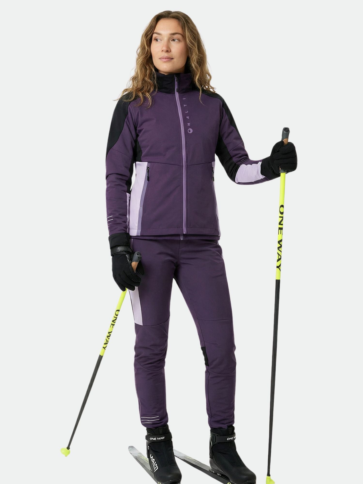 A woman in the Halti Veloce XCT Jacket stands on skis, holding yellow ski poles against a plain background.