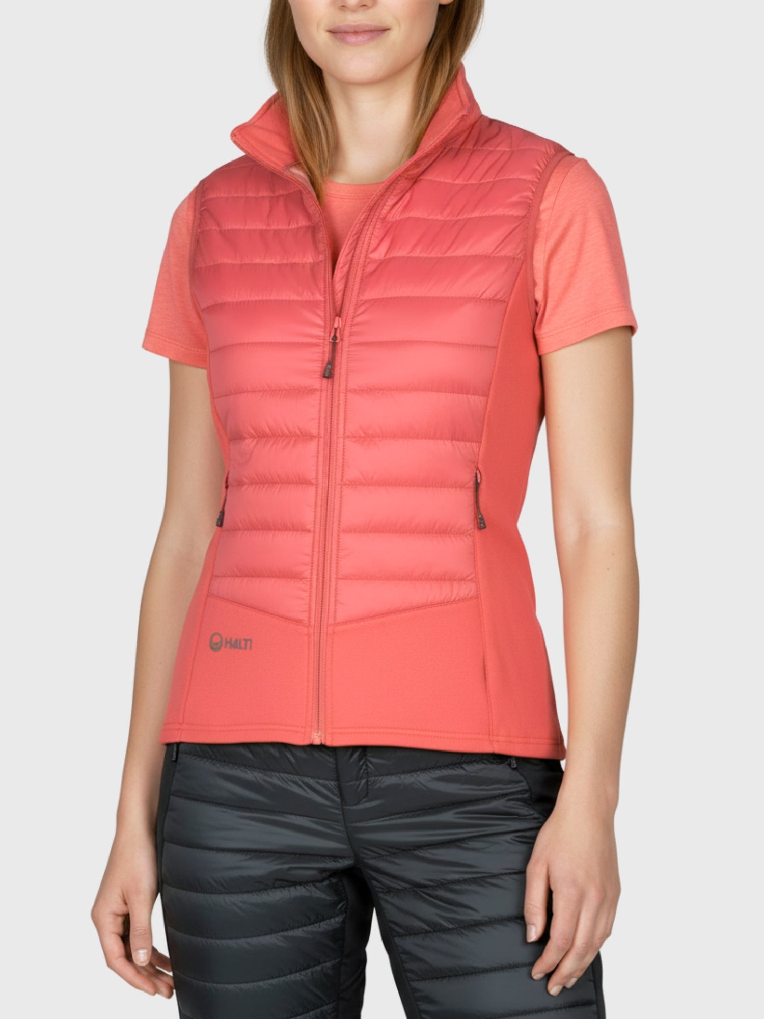 A woman wears the Halti Veidnes Women's Insulation Vest in coral with a shirt and black quilted pants against a plain background.