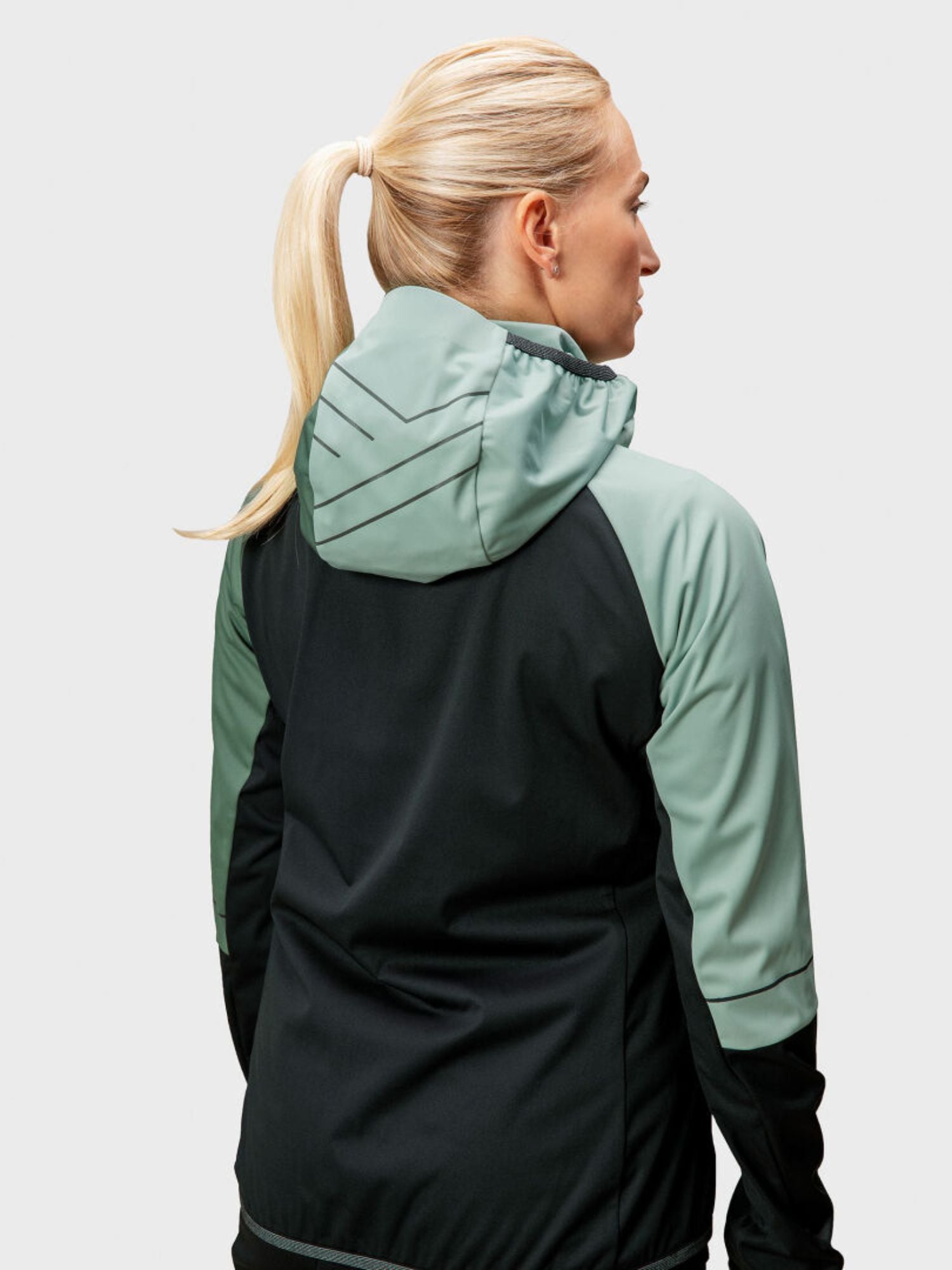 A blonde woman in a ponytail is seen from behind wearing the Halti Exhale Stormwall Jacket Women's in black and light green.