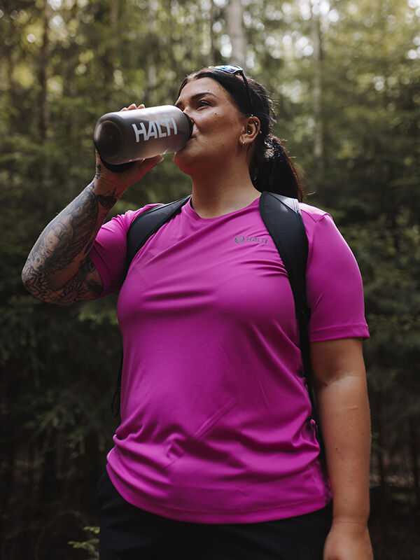 Woman in a bright pink Halti Salves Plus T-shirt drinks from a Halti bottle outdoors among trees.