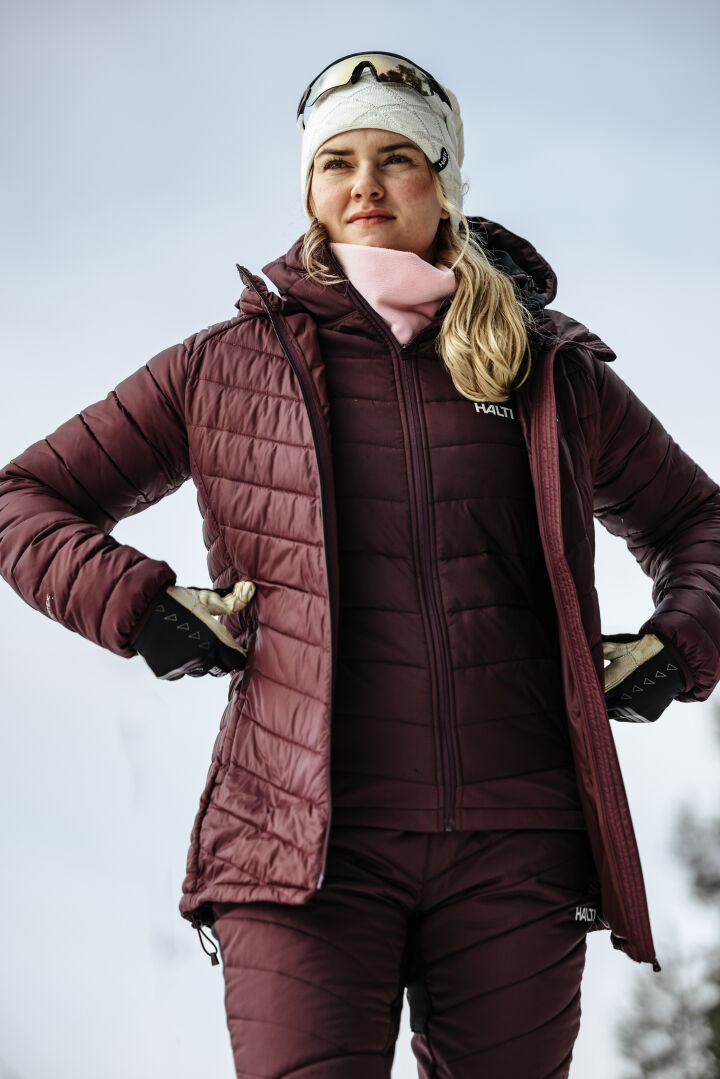 A woman wears the Halti Hanki 2 Hybrid XCT Jacket in maroon, with matching pants, gloves, a white hat, and sunglasses.