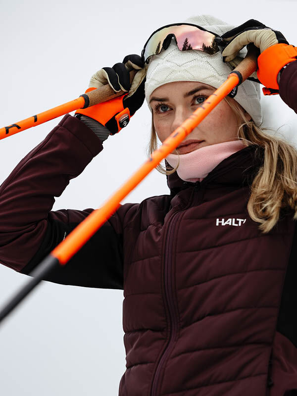 Woman in winter gear adjusts ski goggles, holds orange poles, wears maroon jacket and Halti Crostie Merino Beanie.