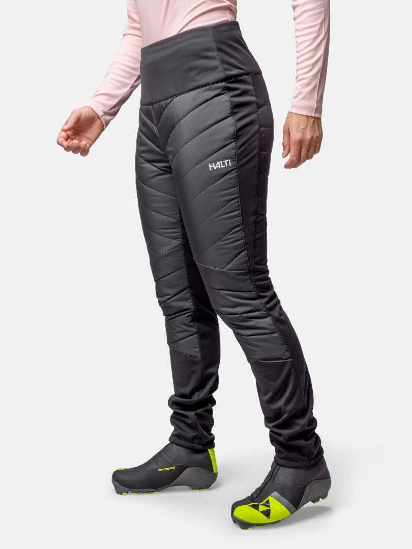 A person wears Halti Hanki 2 Hybrid XCT Pants, a light pink long-sleeve shirt, and black shoes with yellow-green accents.