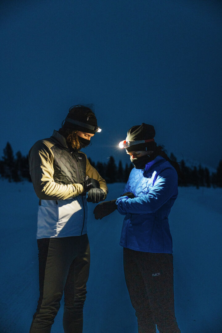 Two people in winter jackets and Halti Framover XCT Pants stand at night, checking watches amid snowy trees.