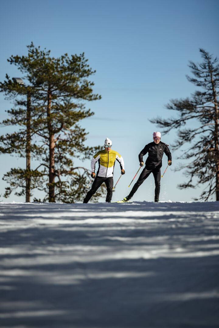 Two people wear Halti Framover XCT Jacket Men's while cross-country skiing on a snowy trail under a blue sky.
