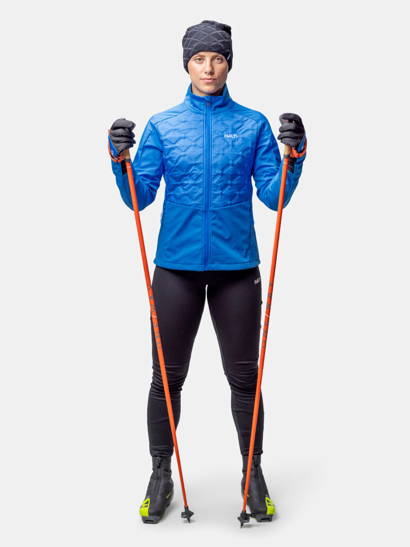 Woman in Halti Framover Hybrid XCT Jacket and black leggings holds orange ski poles against a plain background.
