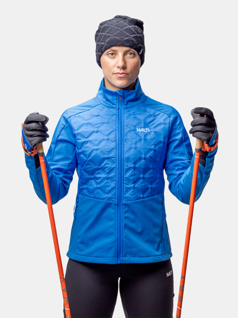 Woman in Halti Framover Hybrid XCT Jacket, black hat, gloves, holding two orange ski poles against a plain background.