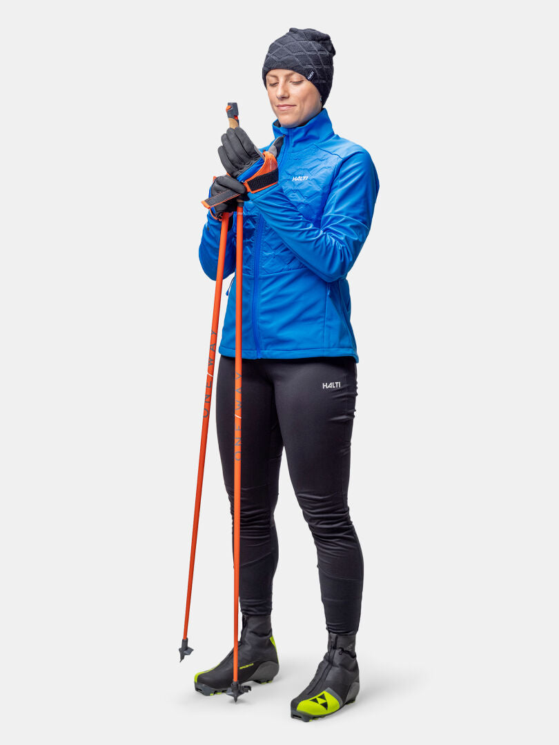 Woman in Halti Framover Hybrid XCT Jacket and hat holding orange ski poles against a plain white background.