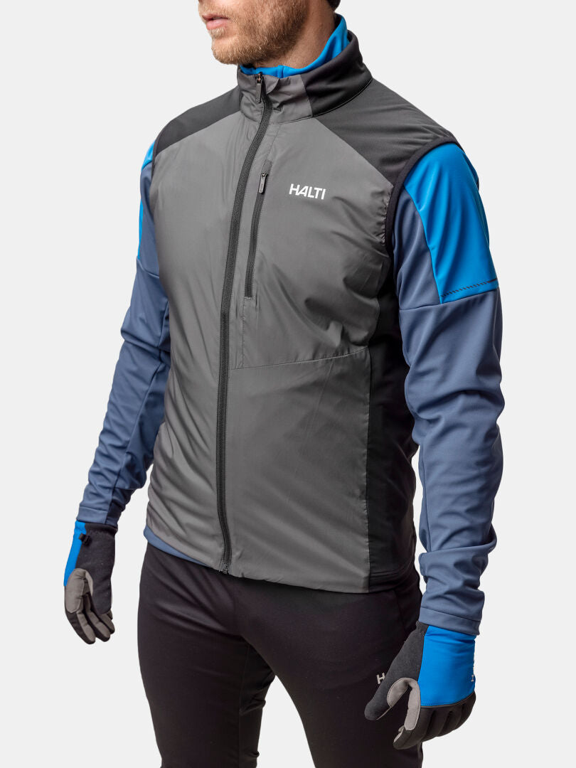 A man wears the Halti Framover XCT Vest Unisex (gray/blue), blue gloves, and black pants on a plain light background.
