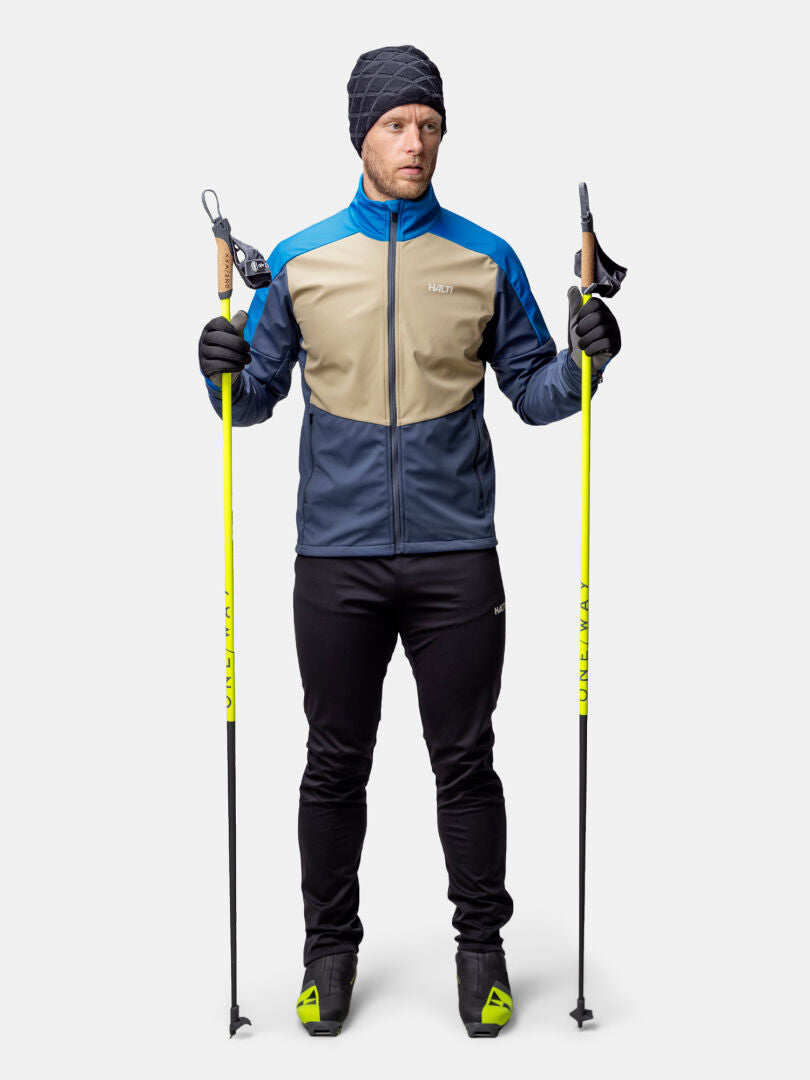 A man wears the Halti Framover XCT Jacket Men's, holding ski poles against a plain background.