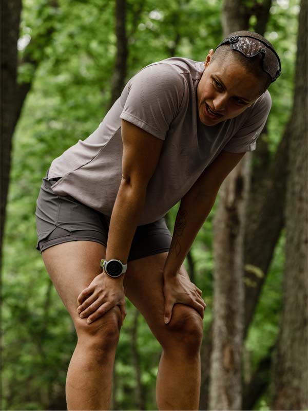 Woman wearing Halti Uurto Shorts rests with hands on knees in a wooded area, catching her breath.