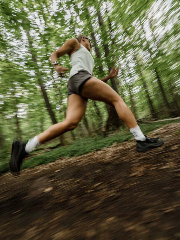 Woman running on a dirt trail in Halti Uurto Shorts, surrounded by trees, captured from a low angle with motion blur.