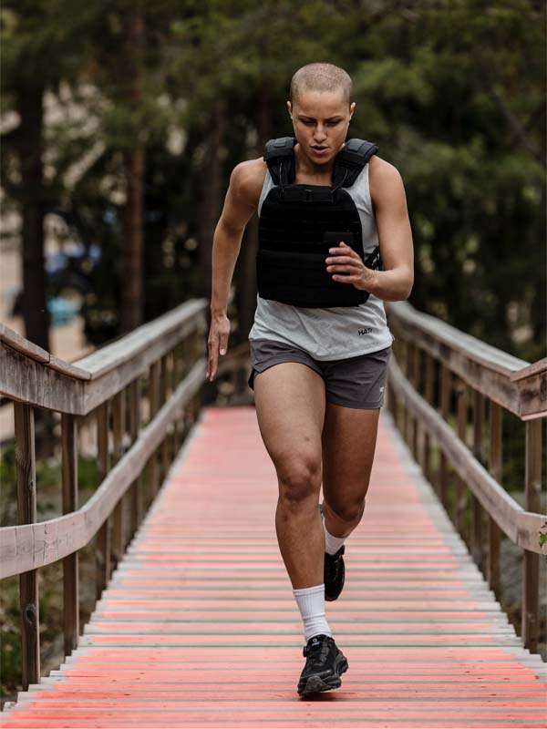 A person with short hair runs up red outdoor stairs, wearing Halti Uurto Shorts Women's, a weighted vest, and black shoes.