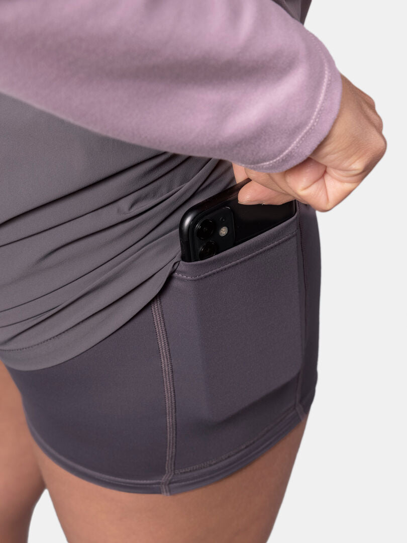 A person slips a black smartphone into the side pocket of Halti Uurto Shorts Women's, wearing a long-sleeve top.