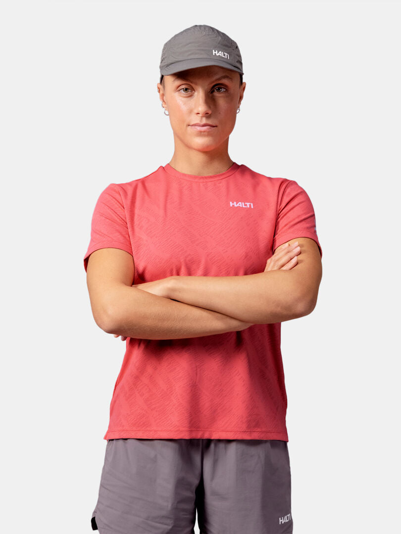 Person in a red Halti Oulanka T-shirt, gray Halti shorts, and cap, standing with arms crossed.