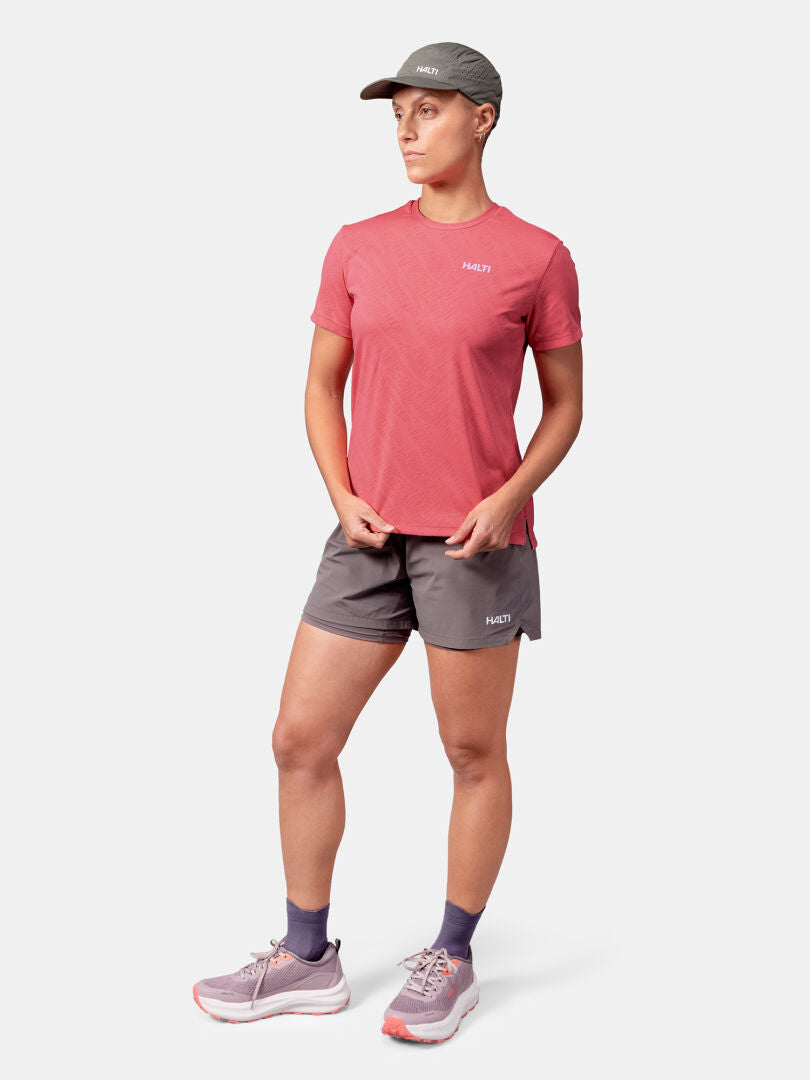 A woman wears a Halti Oulanka T-shirt with gray shorts, purple socks, cap, and sneakers against a white background.