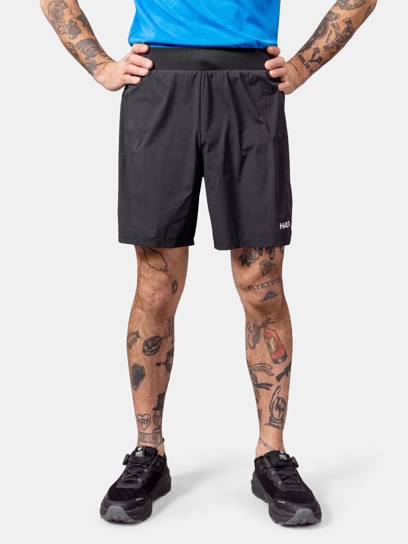 Man with tattooed legs stands hands on hips, wearing Halti Uurto Shorts Men's, black sneakers, and blue shirt.