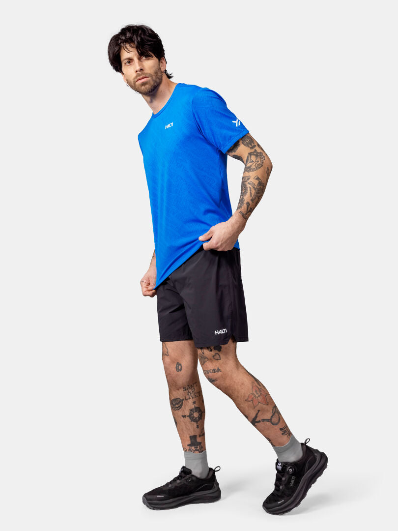 A man wears a blue Halti Oulanka T-shirt, black shorts, and black sneakers against a plain white background.