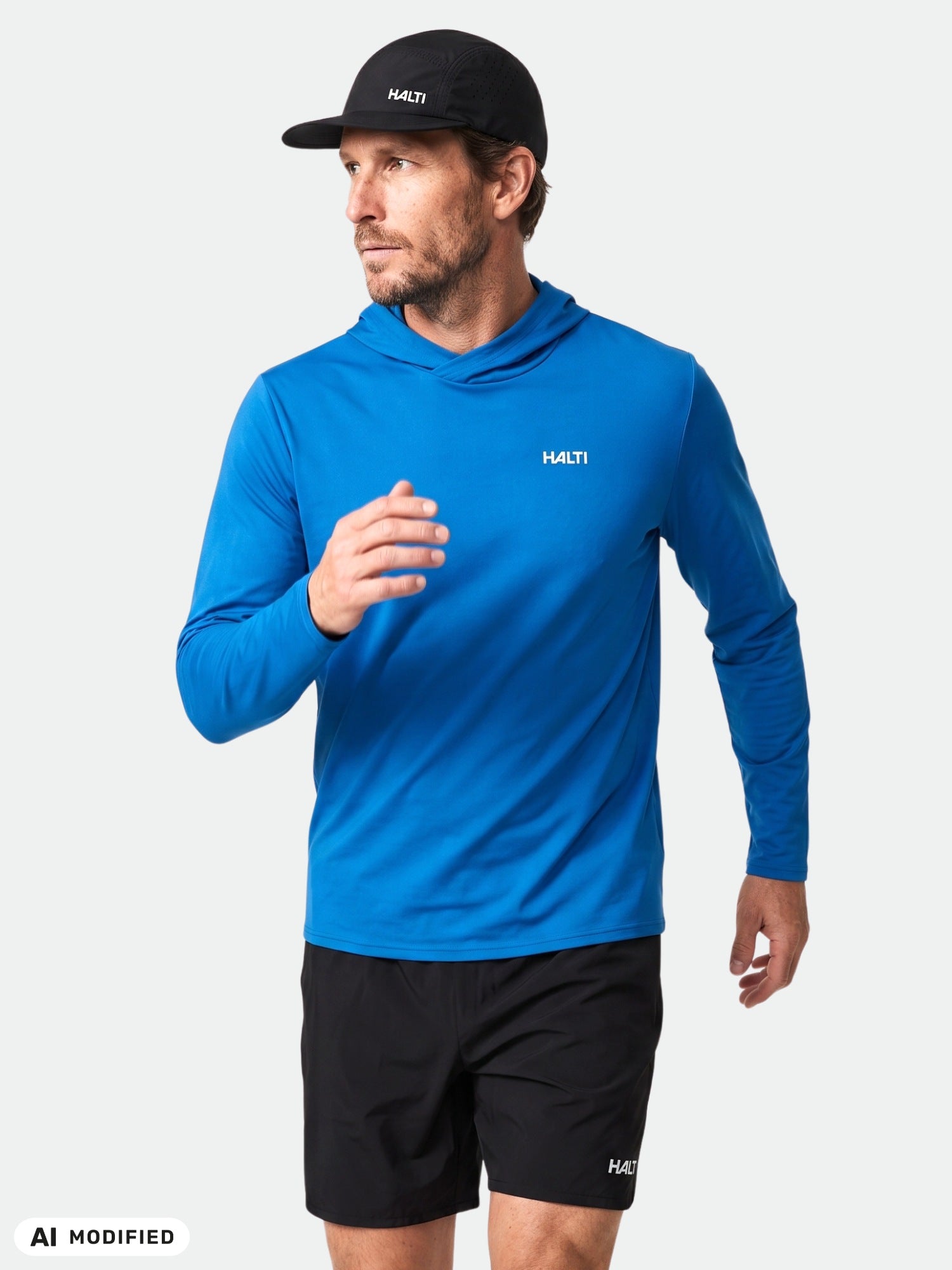 Man in Halti Kaltio Training Hoodie, blue, UPF 50+, with black shorts and cap, looks to side on light background.