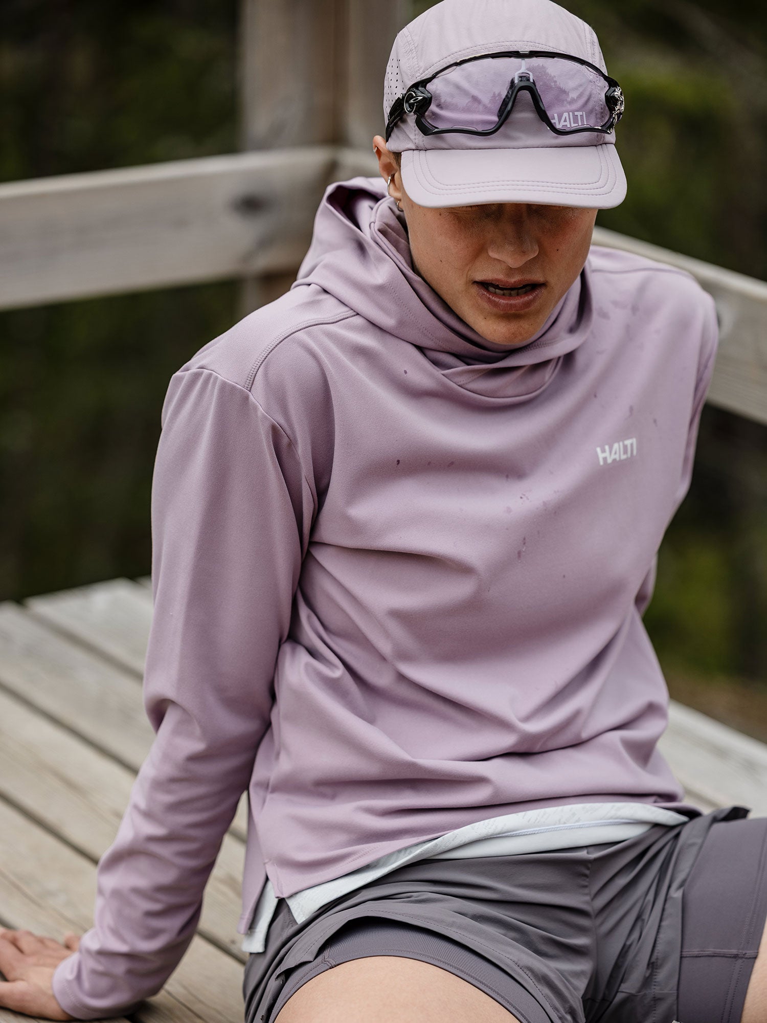 A woman in a Halti Kaltio Training Hoodie, shorts, cap, and sunglasses sits on a wooden bench outdoors.