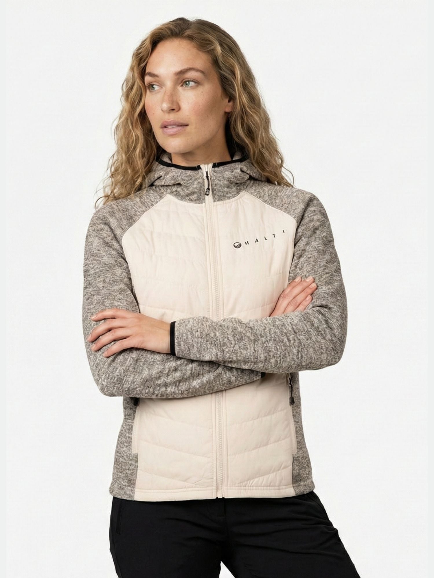 A woman with long wavy hair wears a Halti Streams Hybrid Layer Jacket Women's and stands with her arms crossed.