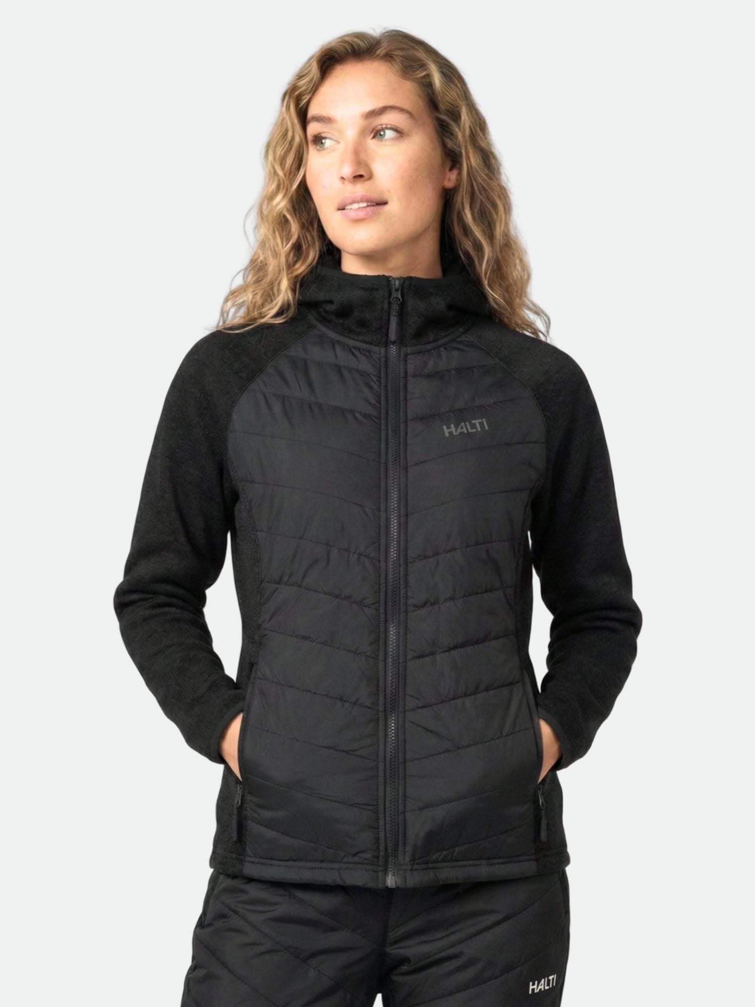 A woman models the Halti Streams Hybrid Layer Jacket Women's in black, hands in pockets, against a light plain background.