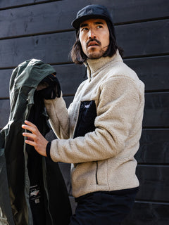 A person in a Halti Lumista Polar Fleece Jacket holds a green windproof jacket by a dark wooden wall.