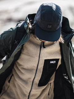 Person wearing a black cap, green windproof jacket, and Halti Lumista Polar Fleece Jacket Mens, looking down.