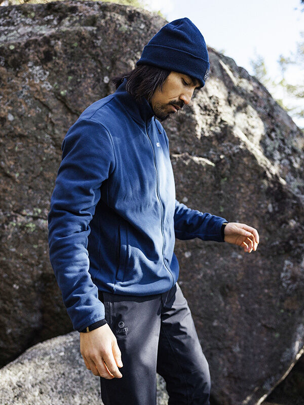 Wearing the Halti Rockmoon Fleece Jacket, a man stands by a large rock, layering for cold outdoor conditions.
