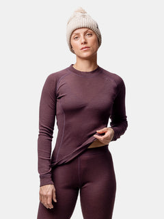A person wears a beige knit hat and the Halti Hossa II Merino Base Layer Set Women's in purple against a plain background.