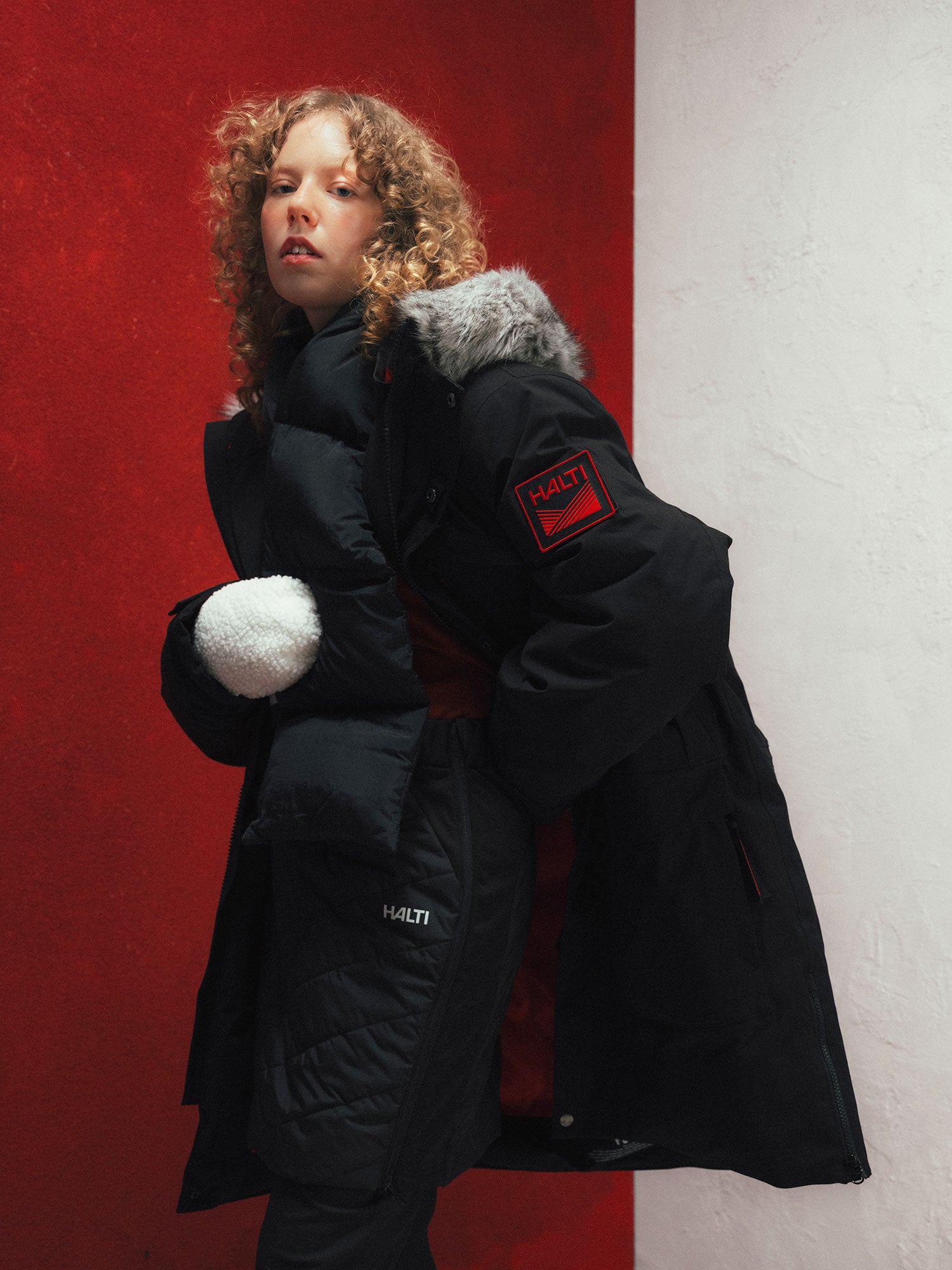 Person with curly hair in a Halti 1976 Down Parka Unisex holds a white ball by a red and white wall.
