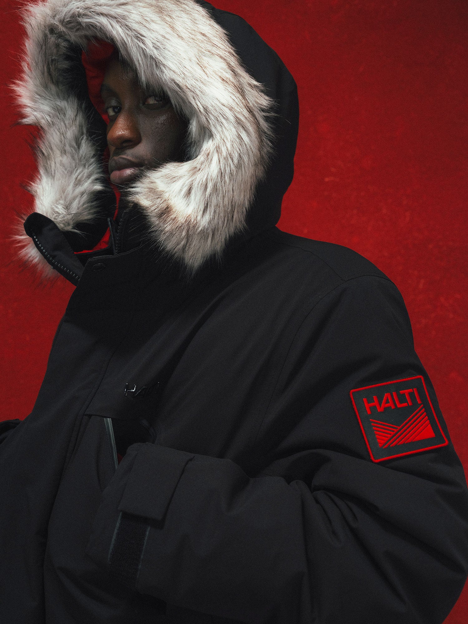 A person in a Halti 1976 Down Parka Unisex with a fur-lined hood stands against a solid red background.