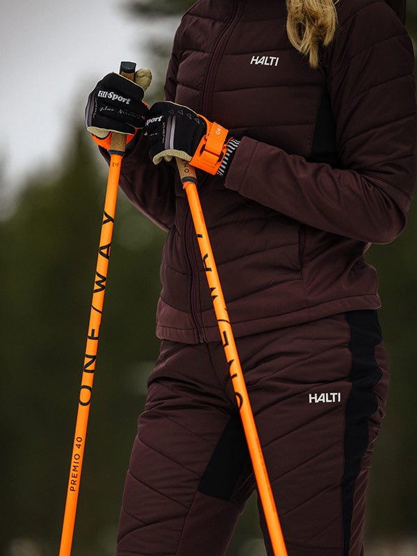 Person wearing a drak red jacket and pants with orange ski poles, against a blurred natural background.