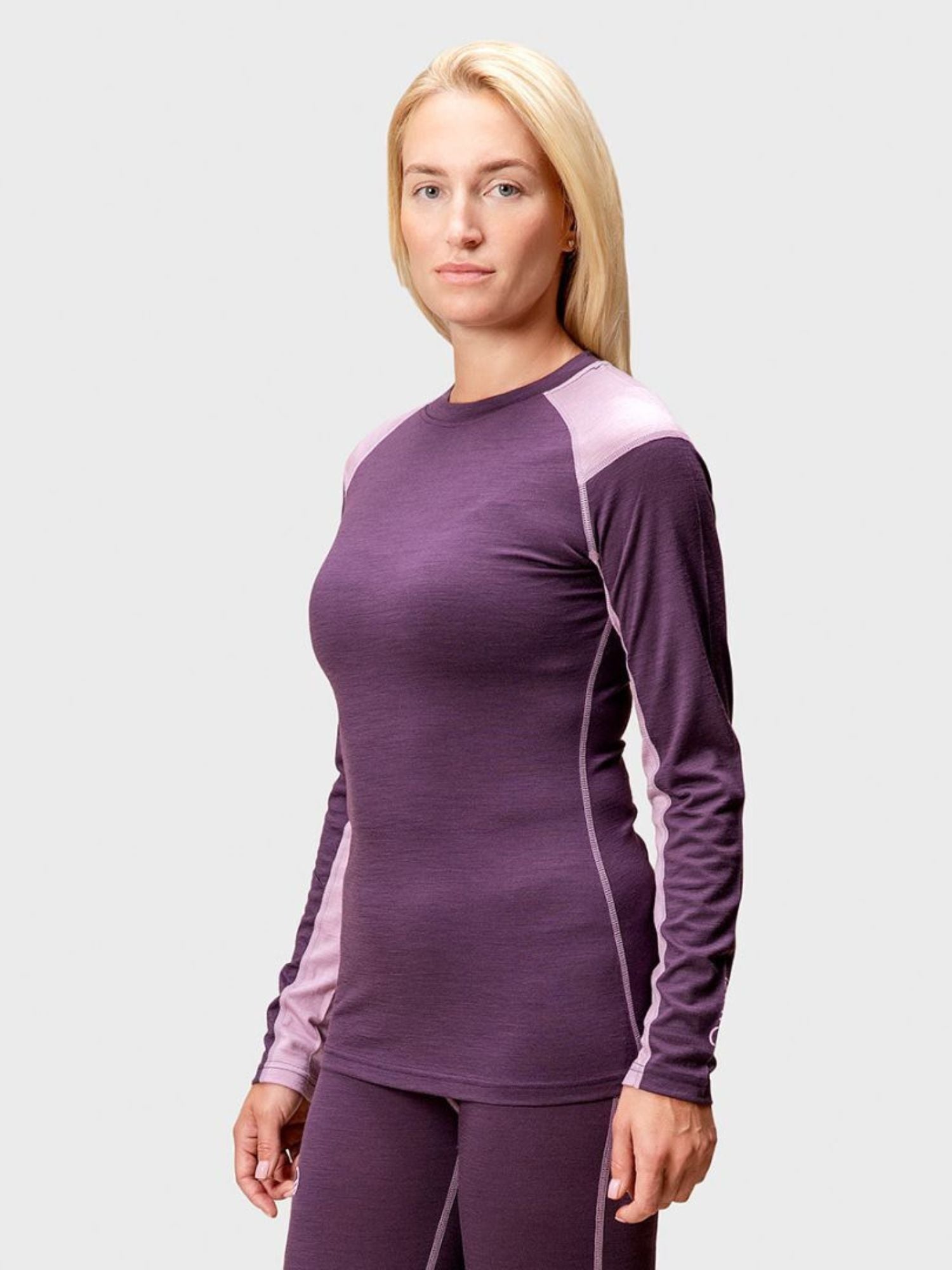 A woman wears the Halti Pihka II Merino Baselayer Shirt in lavender with matching pants, standing against a plain background.